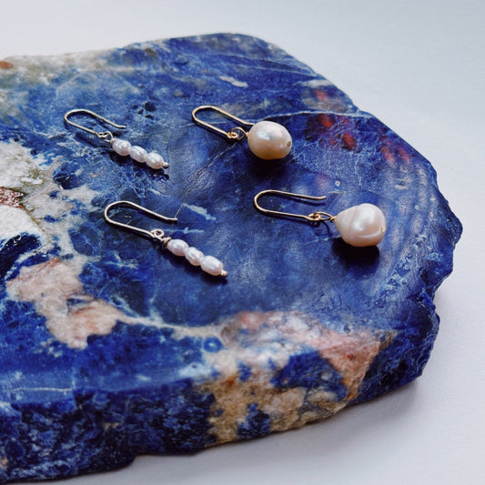 A pair of hook earrings featuring a single seed pearl on a blue stone, with gold shepherd hooks.