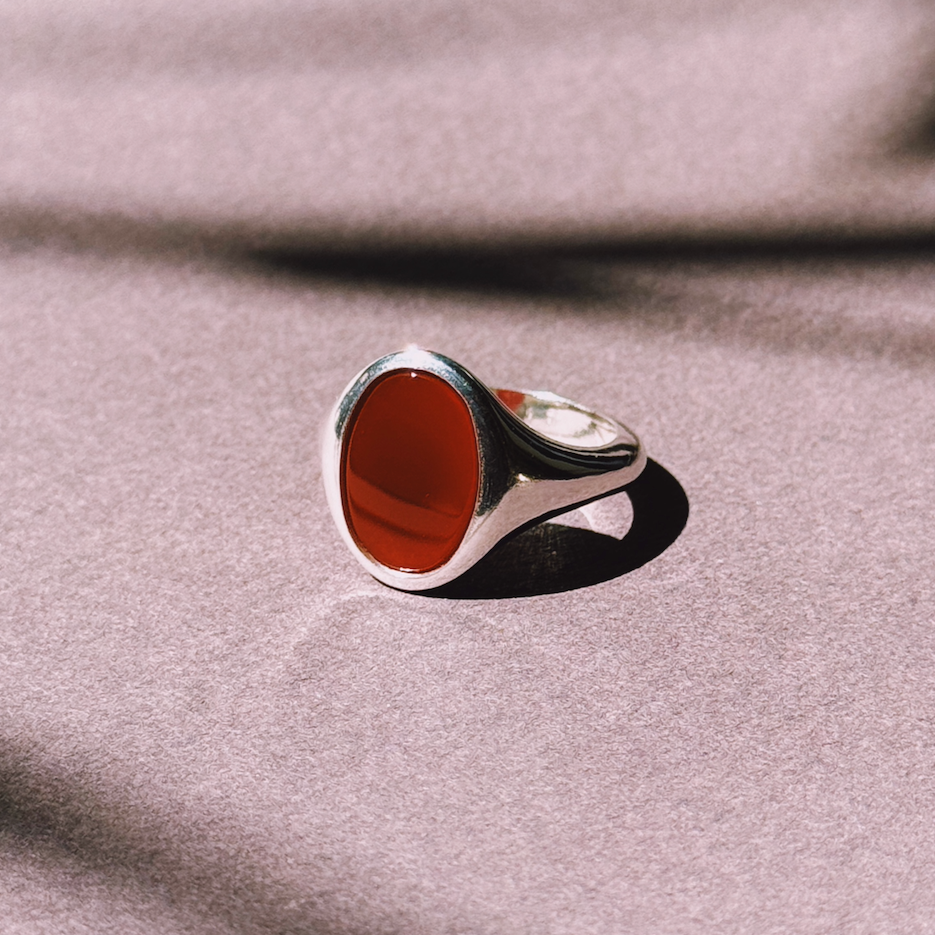 An oval-shaped signet ring with a red Carnelian or Onyx stone set in a silver band, placed on a textured background.