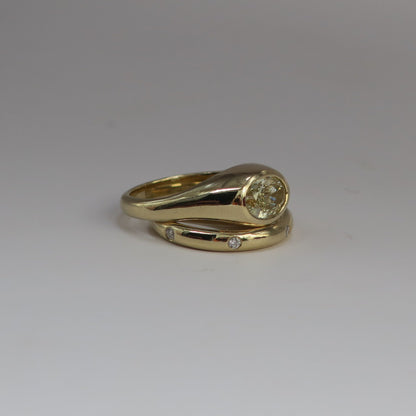 A yellow gold ring with a curved band, featuring an oval diamond in a bezel setting sitting on top of a gold band with  seven small round diamonds flush set along the band, photographed on a neutral gray background.