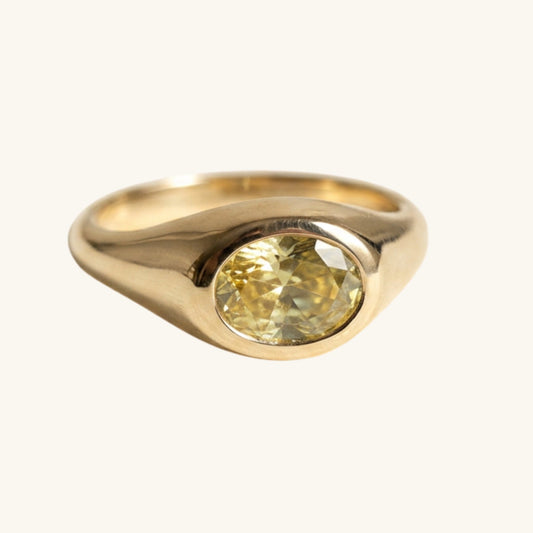 A gold signet ring featuring a large, oval-cut yellow diamond, shown on a clean off-white background.