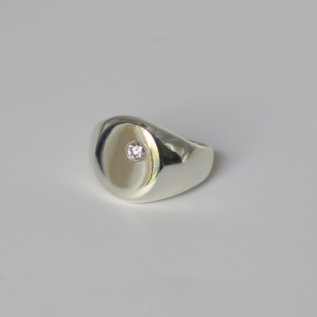A polished silver signet ring with a small round diamond set off center on the round face of the ring. Photographed on a clean light gray studio background.
