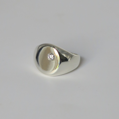 A polished silver signet ring with a small round diamond set off center on the round face of the ring. Photographed on a clean light gray studio background.