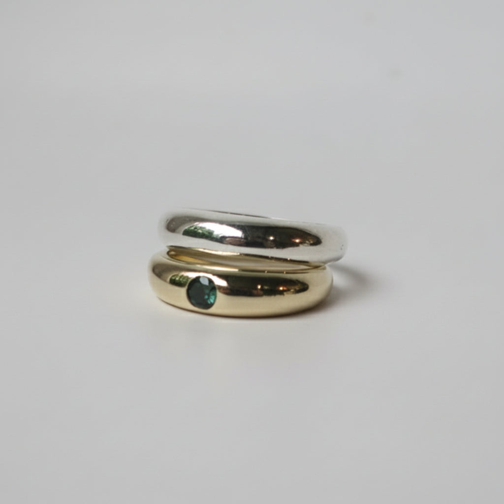 Silver ring stacked ontop of a gold ring with a green gemstone on a light gray background