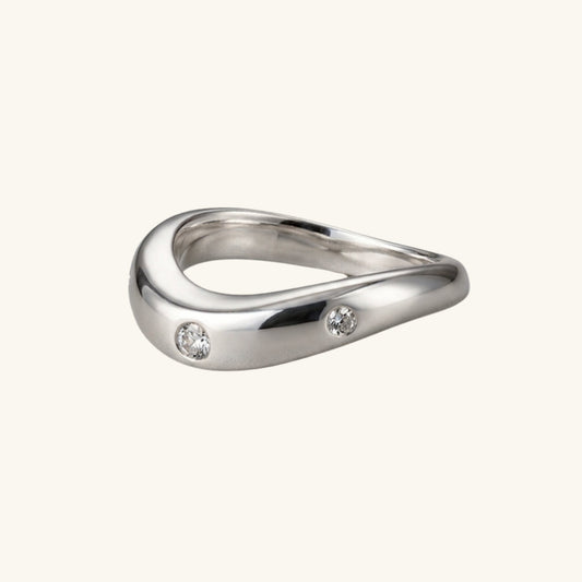 A polished silver ring with a wavy, organic shape and two small clear gemstones, shown on a light cream background.