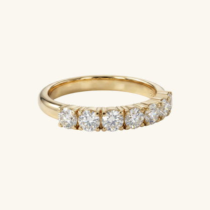A yellow gold eternity ring featuring seven round brilliant-cut diamonds set in a half-channel row, shown on a light cream background.