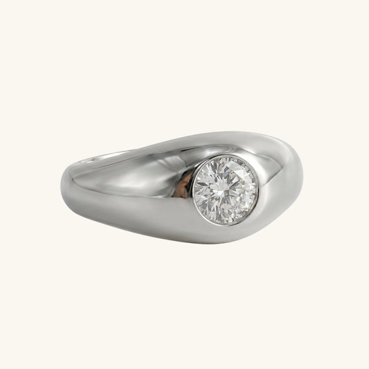 A white gold and diamond chunky ring that's shape is determined by the round diamond in the centre of the band. The ring is sitting on a cream background