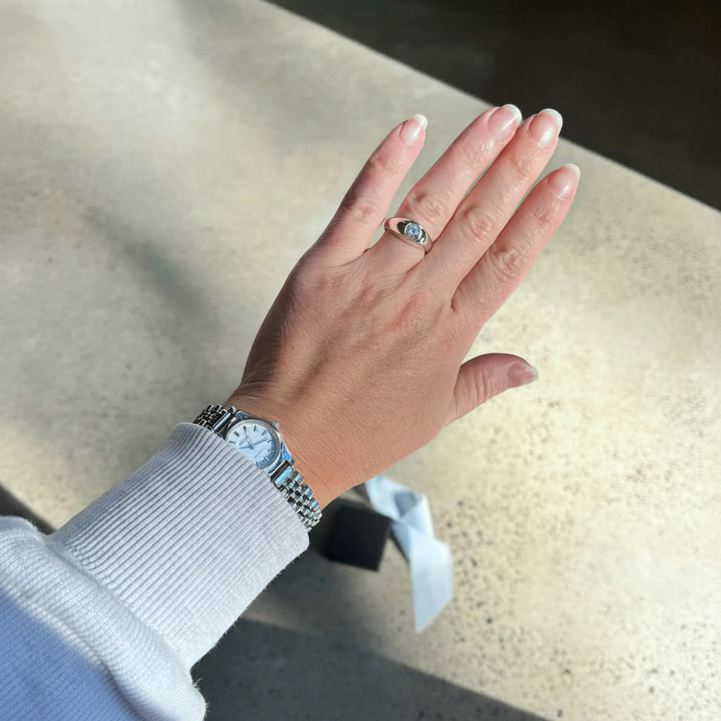 A silver-toned ring with a small oval blue gemstone is worn on the ring finger of a person's hand, who is also wearing a silver-toned watch with a white dial and metal bracelet. The person is wearing a white ribbed long-sleeve top, and the background is a speckled beige stone floor.