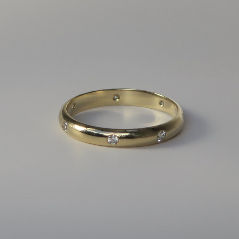 A yellow gold band ring with seven small, evenly spaced clear round-cut diamonds, photographed on a neutral light gray background.