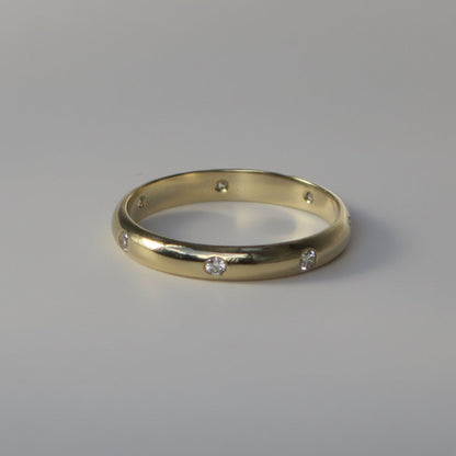 A yellow gold band ring with seven small, evenly spaced clear round-cut diamonds, photographed on a neutral light gray background.