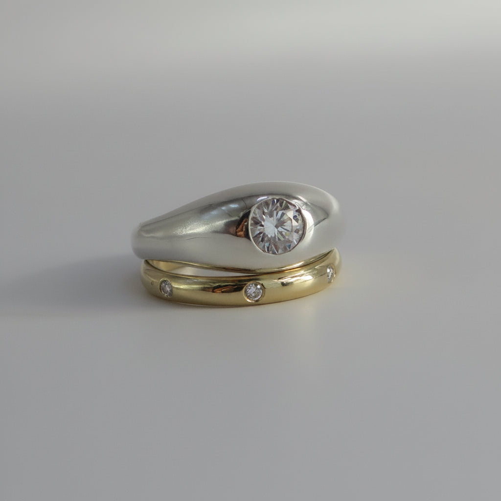 A two-tone wedding ring set featuring a silver band with a round clear gemstone and a yellow gold band with small inset diamonds, displayed on a neutral gray background.