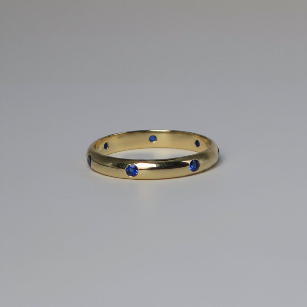 A polished yellow gold band ring featuring seven small, evenly spaced round-cut blue gemstones, photographed on a neutral light gray background.