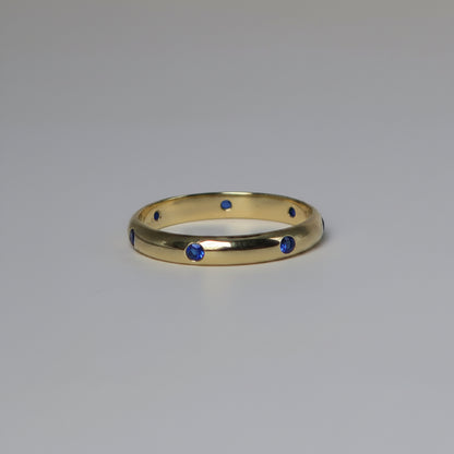 A polished yellow gold band ring featuring seven small, evenly spaced round-cut blue gemstones, photographed on a neutral light gray background.