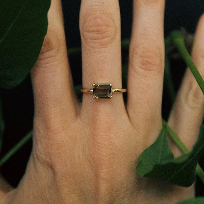 Gold ring with a green gemstone on a person's finger against a leafy background