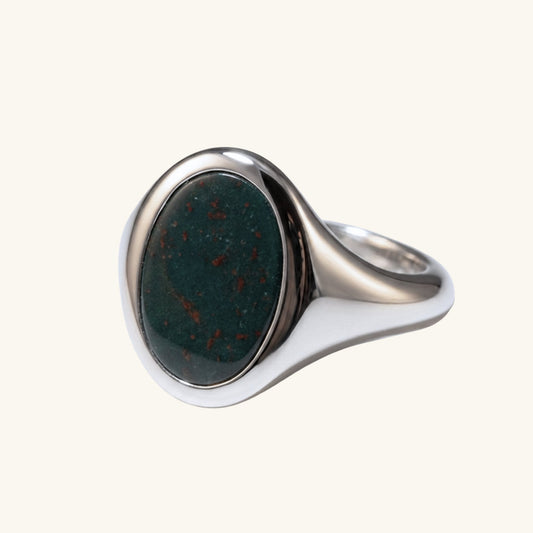A silver signet ring featuring an oval dark green stone with tiny reddish-brown inclusions, set against a solid off-white background.