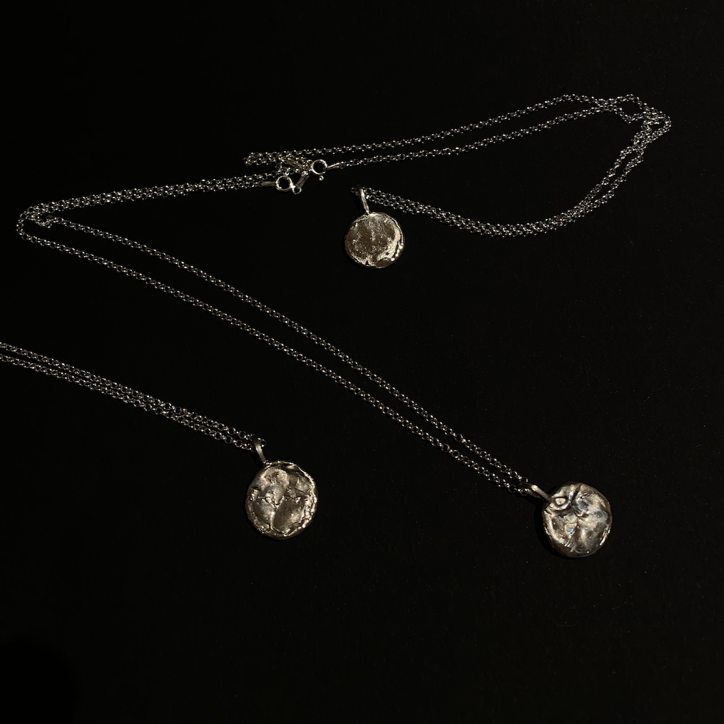 Three necklaces laid out on a dark surface, one with a silver pendant and the other with a gold-plated pendant both attached to respective chains.