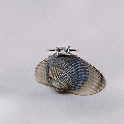 A ring with a square-cut stone, possibly a citrine, smoky, or mint quartz, set in either yellow gold or silver, displayed on a natural seashell.